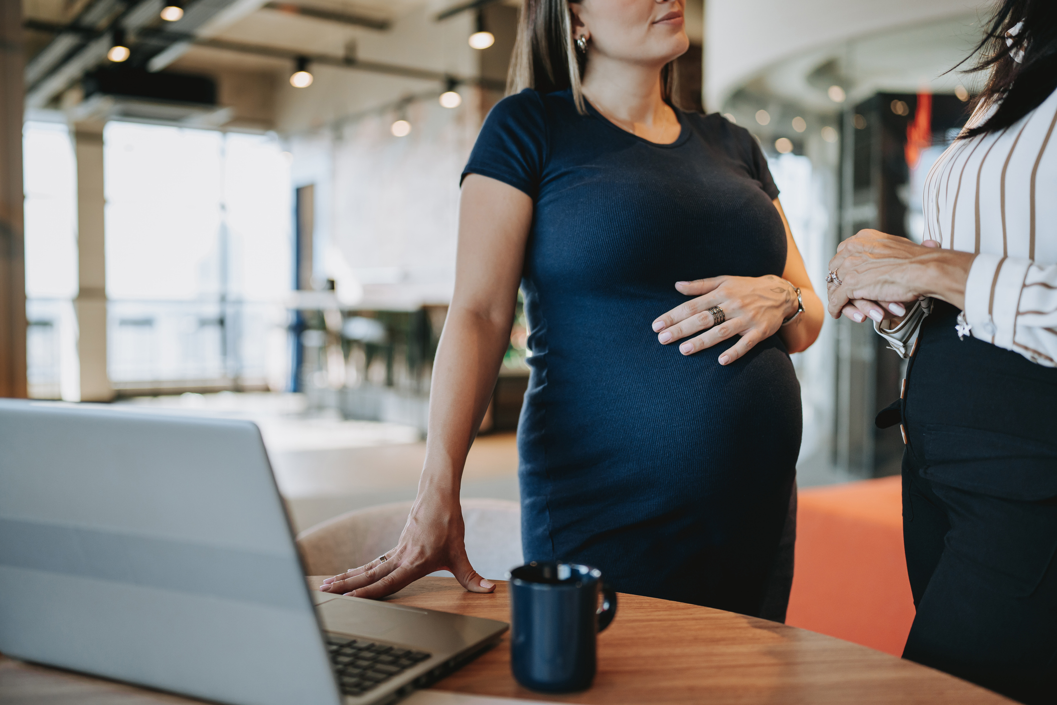 Pregnant woman speaking with another woman at the workplace. Her hand is resting on her belly.